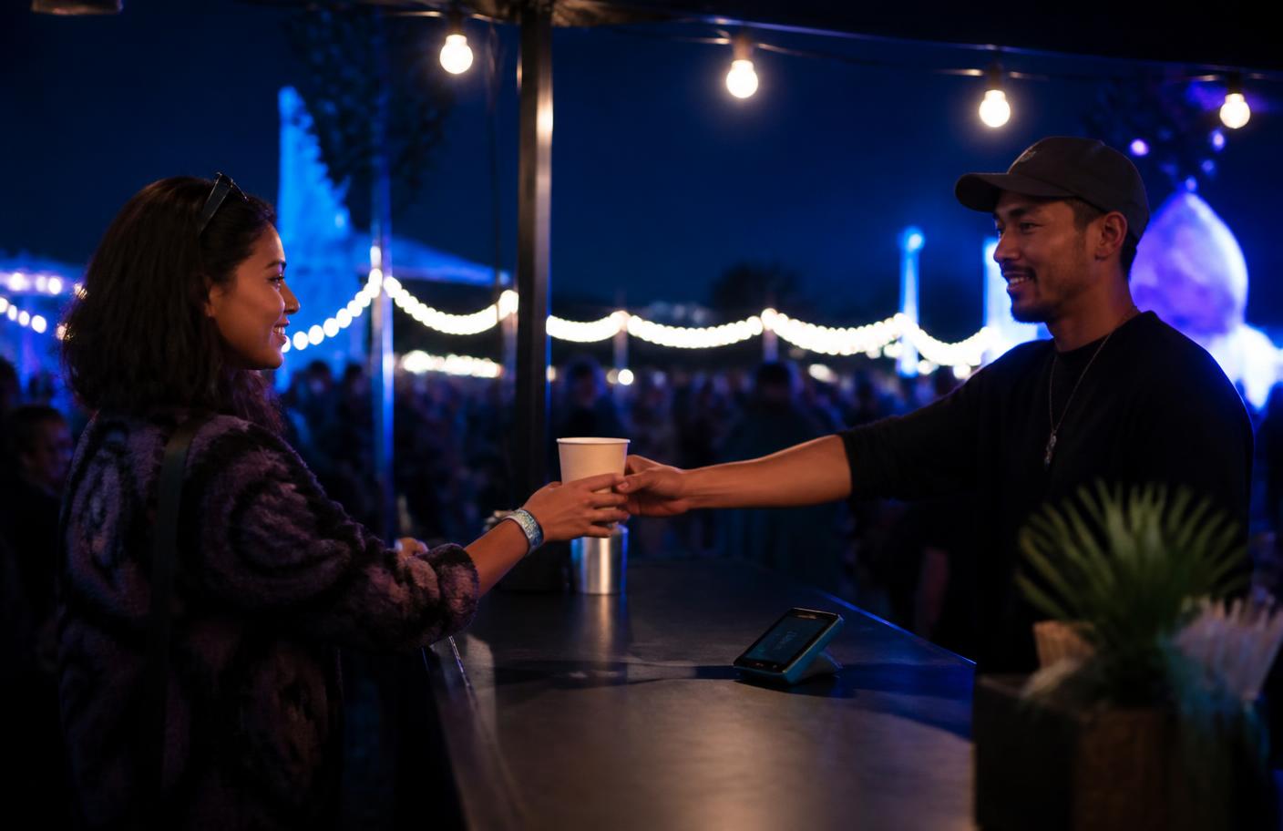 Festival concession vendor serving a customer with a handheld payment terminal on the counter at night